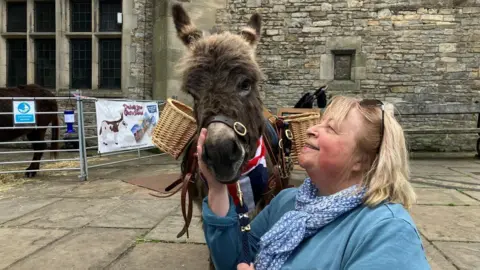 A woman with a short blonde bob, wearing a blue jumper and blue patterned scarf, holds the lead rope of a miniature donkey. The donkey is wearing a union jack neckerchief and a saddle with two baskets on either side. The woman is smiling and petting the donkey's nose. 
