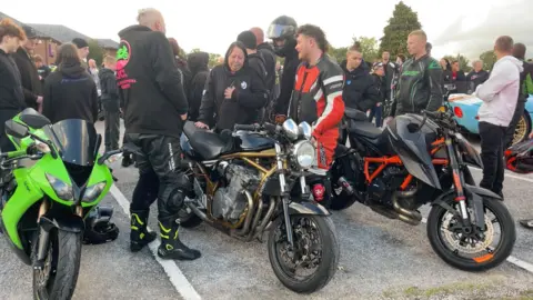 A woman in a black hoodie with long black hair stands beside a gold and black motorbike. She has one hand on the seat and another on her heart. Two men stand either side of the bike, both in biking leathers, one covered by a black hoodie. Behind them are large crowds of people. Two other motorbikes are visible either side of the black and gold bike.