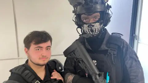 Two young men pose in full military-style cosplay outfits. The man on the left wears a black tactical vest and has short dark hair. The man on the right wears a skull-patterned face mask, night vision goggles mounted on his helmet and carries a large replica rifle.

