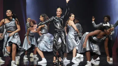 Dancers from The Brit School on stage during a performance. They are all wearing silver outfits, with the exception of the most prominent fiugure, who is dressed all in black. 