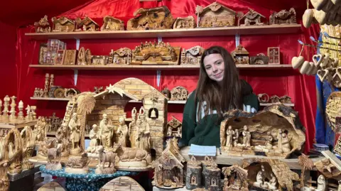 BBC/Kaya Black Hand-carved wood sculptures in a red stall, with a shop worker smiling with long brown hair and wearing a green hoodie.
