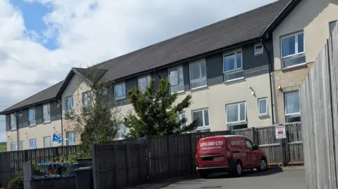 Kingsacre - A modern two-storey apartment building with cream walls and dark cladding, viewed from a fenced parking area. A red service van is parked in the foreground beside wooden fencing, with trees partially obscuring the building under a partly cloudy sky.