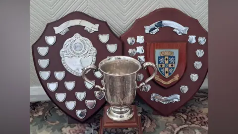Supplied Two shields are propped up side by side with a trophy in front. They are made of dark brown wood and have multiple smaller silver shields on them. The silver trophy has two handles and is mounted on a wooden plinth. 