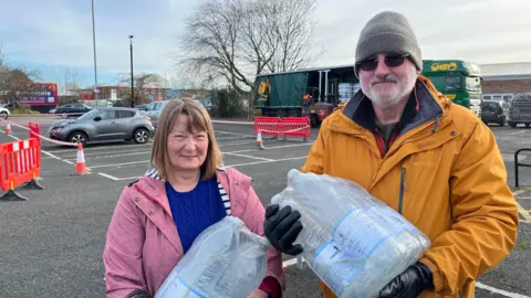 A woman in a pink coat and a man in a yellow coat stand in a car park holding bottled water.