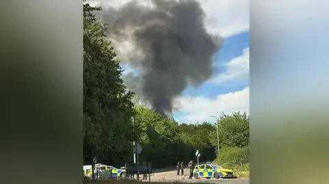 Herts Fire and Rescue Large plume of black smoke rises from behind trees, in the foreground are a number of police cars