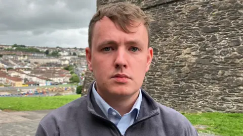 Sinn Féin assembly member Pádraig Delargy is in a grey fleece and blue shirt. 
He is standing beside Derry's historic walls. A number of homes can be seen in the background.