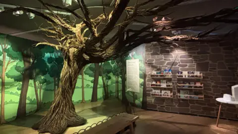 The picture shows a dimly lit room, with a huge depiction of a hawthorn tree. It's intricately woven from wicker branches. Its trunk reaches to the roof where its branches span across the ceiling. Behind it, under green lighting, is a picture of a wooded area. to its right are some bookshelves. Under the hawthorn tree there is a bench. 