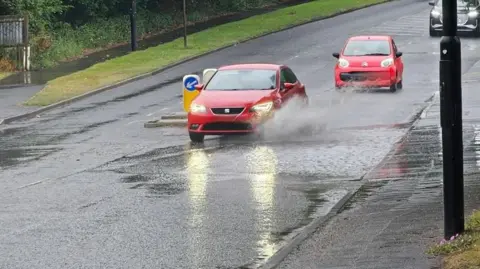 BBC Weather Watchers/Figaro Two red cars drive on a rainy road with large puddles and a grassy verge to the left.