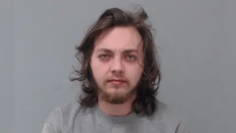 Police Service of Northern Ireland A mugshot of Kornelijus Bracas. He has long, dark hair and a beard. He is in a grey, marled crewneck jumper and is staring at the camera with a neutral expression.