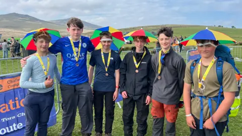 Six boys stand in a line on the moors. They are wearing multi-coloured umbrella hats with medals around their neck. The moors are visible behind them.