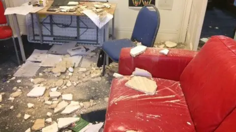Ison Harrison Parts of a ceiling, dropped on a floor, showing a red sofa, large parts of plaster, a blue chair, a desk and paperwork.