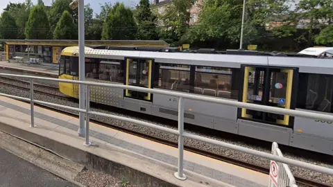 A yellow-and-grey tram arriving at Didsbury Village Metrolink stop on a clear day. 