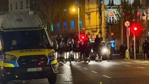 Several police officers on horses enter a city street passing a Garda (police) van. It is dark - clearly nighttime - and two traffic lights to the right of the image are on red. 