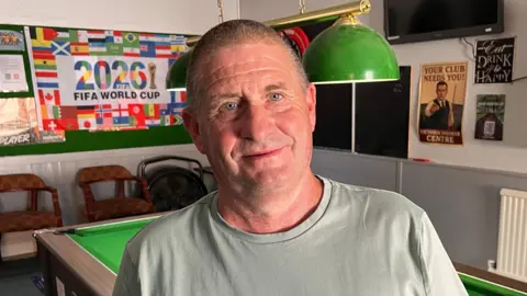 BBC/Phil Harrison Victoria Snooker Club owner Graham Martin stands in front of a pool table with a 2026 FIFA World Cup poster in the background