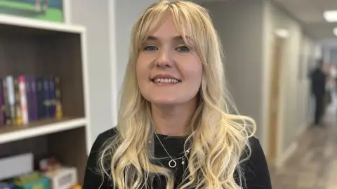 Emma Baugh/BBC Sadie, a woman with shoulder-length golden hair, wearing a black T-shirt, smiling for the camera.