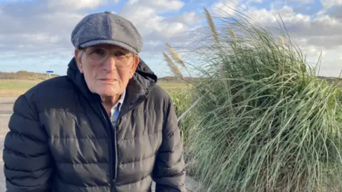 An elderly man wearing a cap and glasses with a black jacket looking directly into the camera. On the right is a green read bush. 