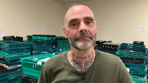 John Ward standing in front of stacks of empty food boxes. He has short salt and pepper hair and trimmed beard and blue eyes. He is wearing a green t-shirt and has a neck tattoo.