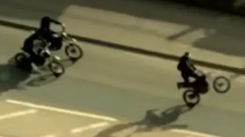 Three people on bikes, in a road, the image is taken from a plane so slightly blurred. One person looks like they are doing a wheelie. 