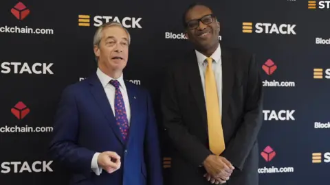 Reform UK leader Nigel Farage (L) stands with former Chancellor Kwasi Kwarteng (R) as they promote cryptocurrency. Farage wears a dark blue suit, a white shirt and a blue tie with red blossoms. Kwarteng wears a black suit, a white shirt and a butter yellow tie.