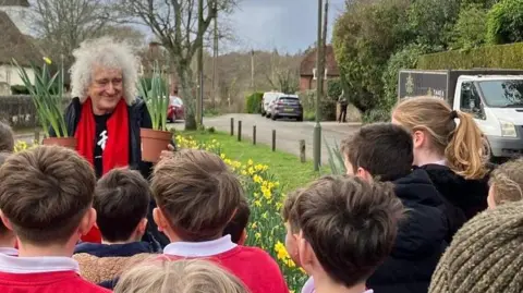Sir Brian May holds two pots of daffodils as he stands with a group of children next to a village green which is covered in daffodils. The viullage church is visible behind him