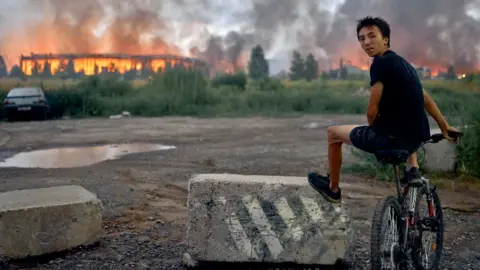  A teenager sits on a bicycle watching flames rise from a burning logistics warehouse following a Russian ballistic missile attack on Kyiv and the surrounding region on August 5, 2026 in Kyiv, Ukraine. 