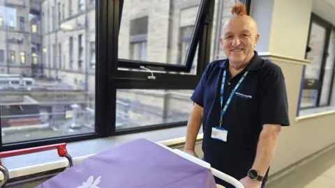 Aisha Iqbal/BBC A middle aged to slightly older man stands smiling. He is bald but wears some Christmas tinsel on his head in afaux Mohican style. He is smiling. He stands in a corridor gripping a hospital trolley. He wears a dark blue t shirt and a lanyard/badge which identify him as a staff member at Bradford Teaching Hospitals.