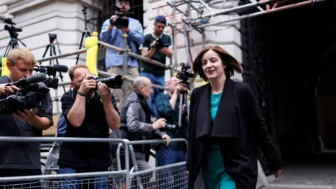 PA Media Varios fotógrafos toman fotos de Bridget Phillipson a su llegada a Downing Street. Se encuentran a su derecha, detrás de una valla metálica. Phillipson tiene el pelo castaño hasta los hombros y lleva un abrigo oscuro y un vestido turquesa.