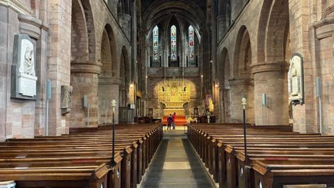 The inside of Shrewsbury Abbey with rows of pews either side of the centre aisle. In the background, you can see the altar and stained glass windows