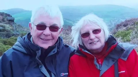 Family photo Head and shoulders photo of Eric and Val in happier times, both smiling to the camera. They both have grey hair and are wearing sunglasses and waterproof jackets. They are on a hill with more hills seen in the background. 