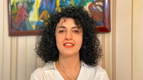 A headshot of Narges Mohammadi  - she looks towards the camera and smiles while wearing a white blouse.