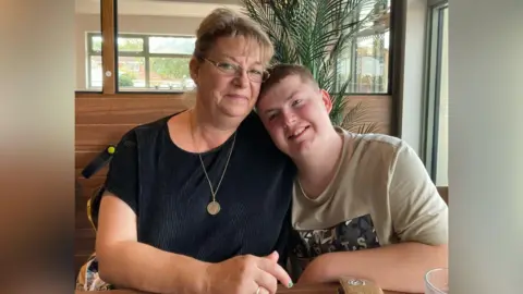 Contact A blonde woman wearing glasses and a black shirt and gold necklace, is sitting next to a teenager wearing a beige colour t-shirt. 