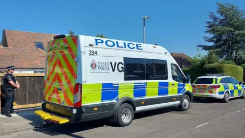 A police car and van parked on a residential road, which has a large tree, bush and house on it. A police officer guards the entrance to a property.