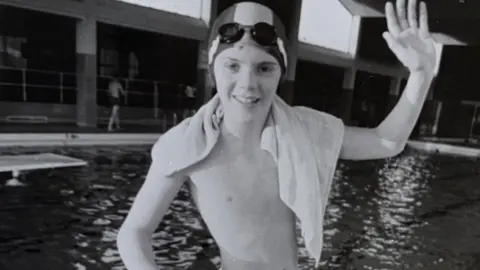 Black and white photograph of a boy standing next to an indoor swimming pool with a swimming cap and goggles on his head and a towel over his shoulders