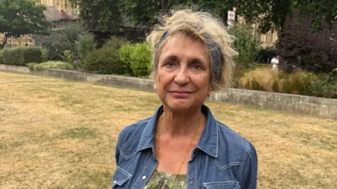 Veronica Dewan standing on a grassy area outside Parliament. She has a blue denim jacket on and a blue headband over white blonde hair.