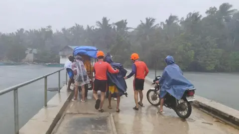 EPA/Shutterstock Two men in red shirts and shorts help a person in a raincoat along a bridge. The sky is grey beyond the bridge and palm trees are windswept.