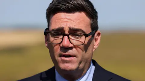 Andy Burnham looking thoughtful against backdrop of a field. He is wearing dark rimmed spectacles
