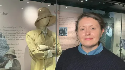 Laura Howarth smiles at the camera in front of the new exhibit. She has brown hair that has been tied back. She wears a black jumper with a denim shirt underneath. 