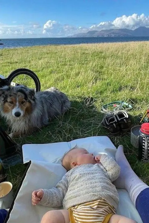 Morag Skelton Baby Hamish lying on a mat on grass, with the coastline and mountains in the background. The family's dog is also sitting in the background.