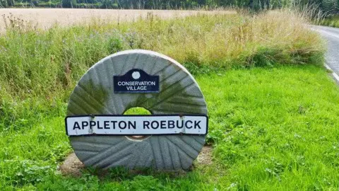 A stone circle with a hole in the middle on a grass verge with the words Appleton Roebuck written on it.