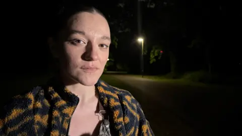 A woman stood looking at the camera with a street light in the background which is next to a green bush.