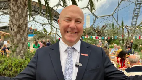 Andy Jasper is wearing a navy suit, white shirt and a blue tie. He is standing in one of the biomes at the Eden project as people enjoy afternoon tea on tables behind him.