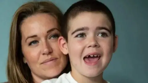 A woman on the left with brown hair behind a boy with his mouth open. A light blue background is behind them.
