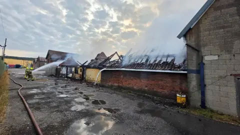 Nottinghamshire Fire and Rescue Service A damaged building with smoke blowing into the air. The roof is missing from the building and firefighters are spraying water into the building. 