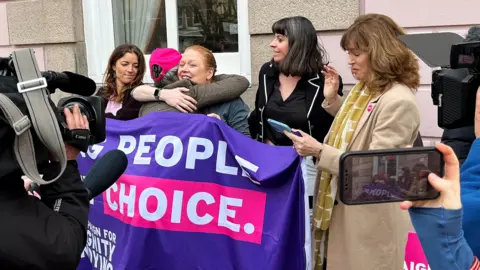 BBC Five women celebrate with hugs next to a banner saying 'people, choice' with news cameras around them, outside on the street in Jersey on Thursday.