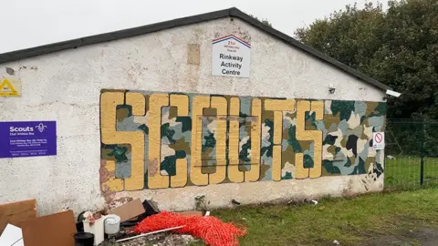 The exterior of the scout hut. The single-storey building is painted white with a yellow and green mural on it that reads: "SCOUTS"