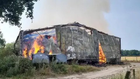 A large barn engulfed in flames with smoke rising in the early morning sky
