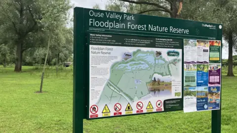 Amy Holmes/BBC A park information board for “Ouse Valley Park – Floodplain Forest Nature Reserve” stands among trees and grass, showing a map, safety warnings, and notices.