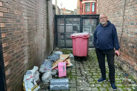 Councillor Alan Gibbons stands in an alley way in his north Liverpool ward. There is rubbish including empty plastic boxes, dog food bags and cardboard strewn over the cobbles. 