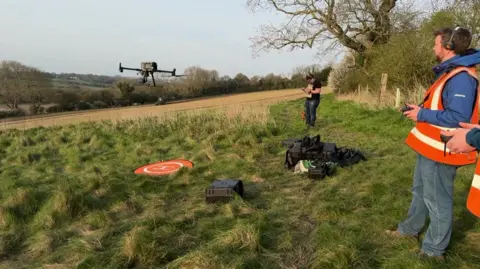 Three people are stood in a field. One of them, in an orange hi-viz vest, is holding controls to a drone that can be seen hovering in the air in front of him.