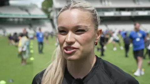A young woman with blonde hair tied back in a ponytail stands on the pitch at Lords.. Children are playing in the background. She is wearing a black t-shirt.